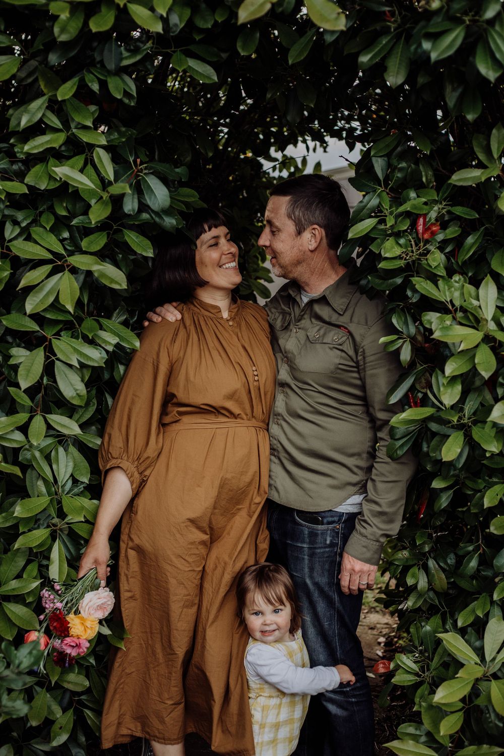 A family embraces among lush green foliage while holding freshly picked flowers.