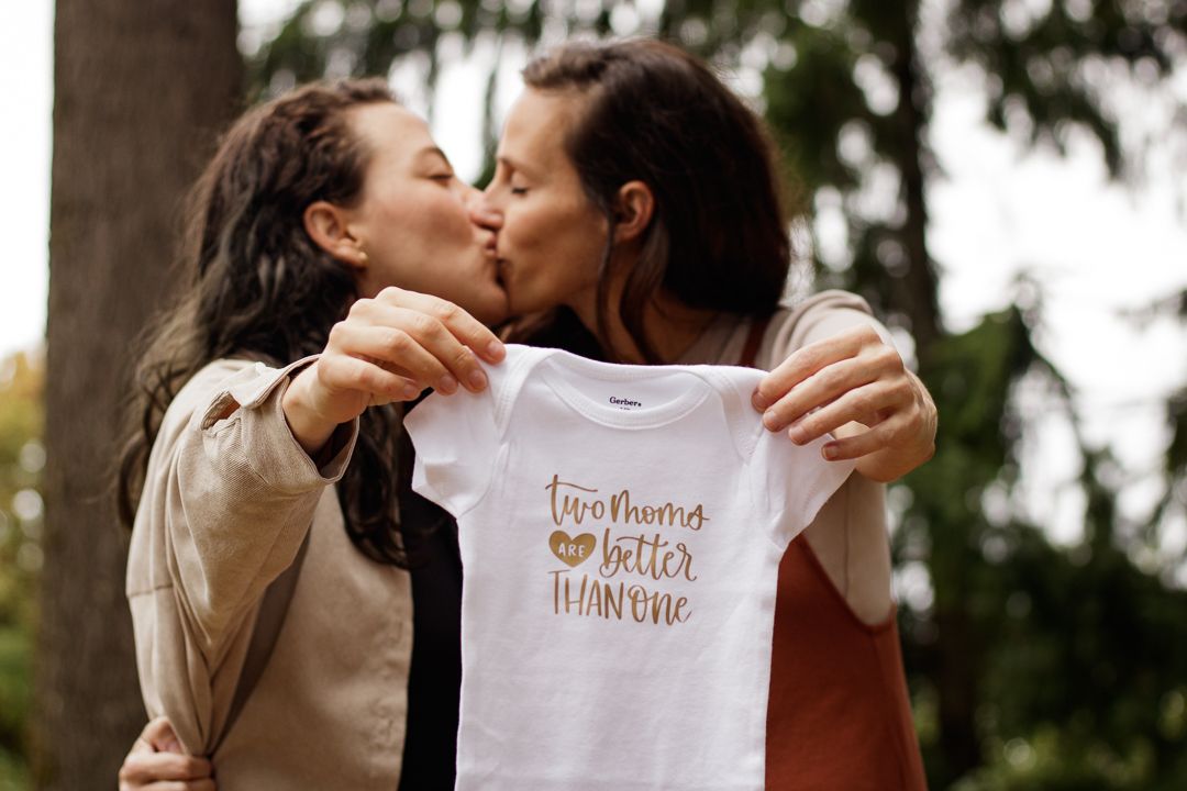 Two people hold up a baby onesie reading "two moms are better than one" in a joyful outdoor setting, surrounded by trees.