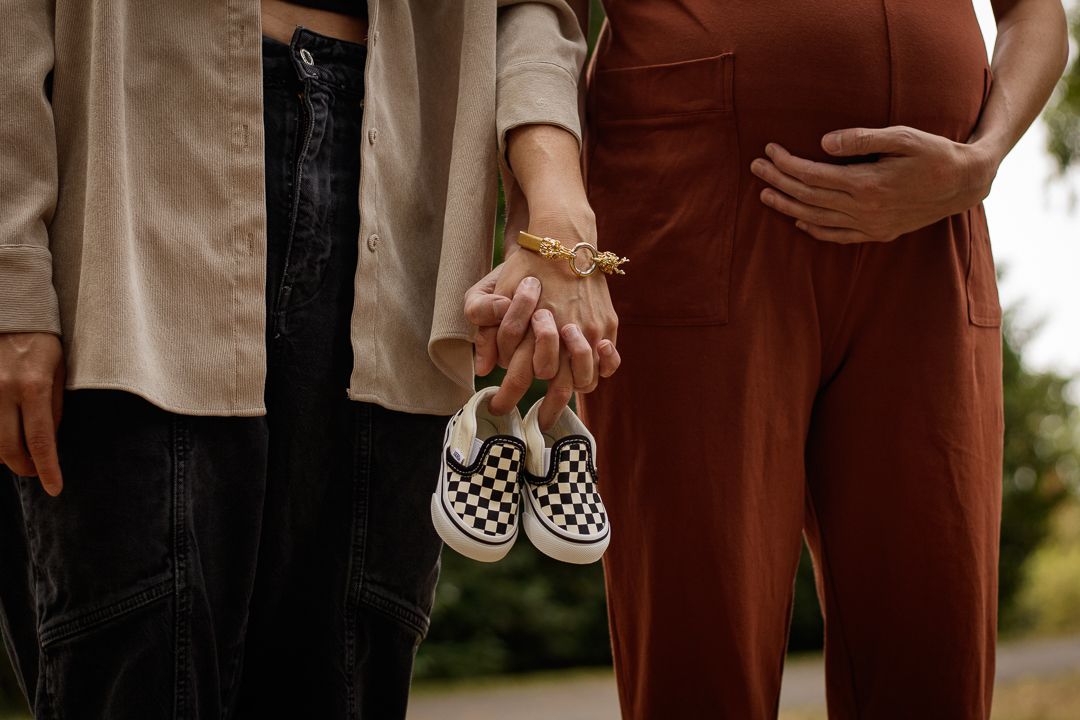 A couple stands closely together holding hands, displaying tiny baby shoes, set against a picturesque backdrop of autumn leaves.