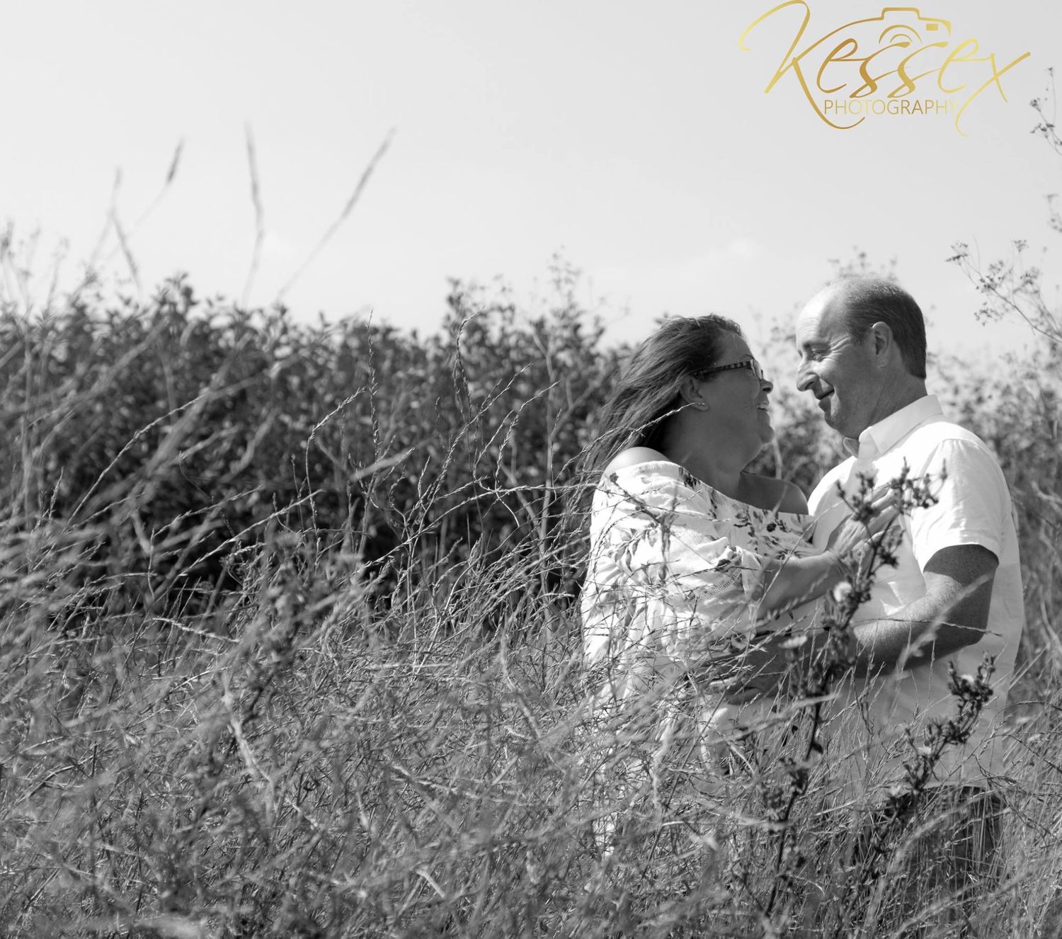 A black and white engagement photo of a couple embracing in tall grass.