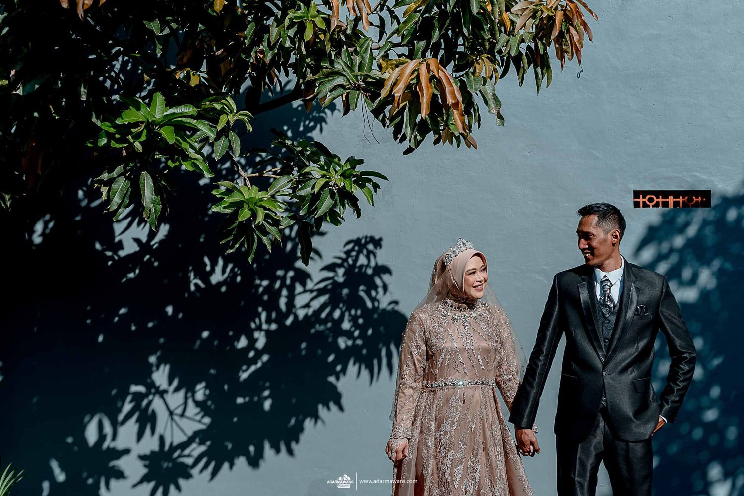 A couple holding hands walks outdoors beneath lush green trees.