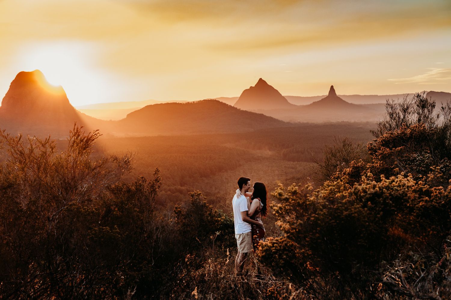 Two figures embrace while standing on a mountain summit during a golden sunset.