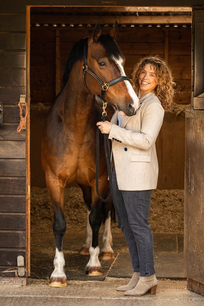 A woman in a beige blazer stands with her horse in a rustic wooden stable setting.