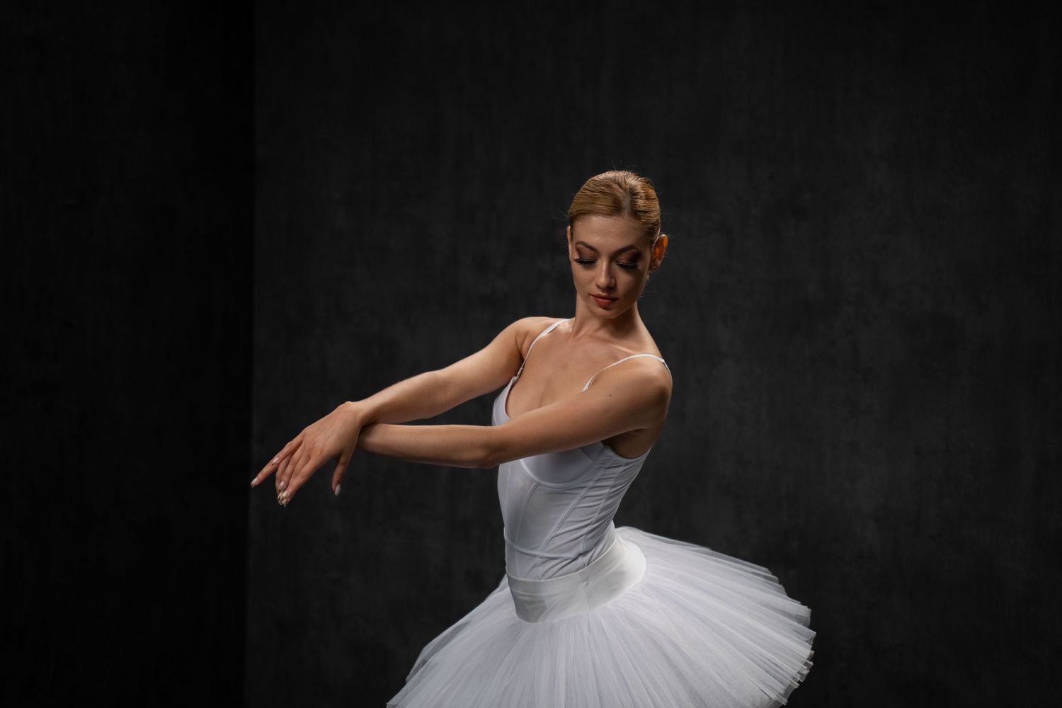 Dancer in classical white tutu performs graceful movements in dark studio.