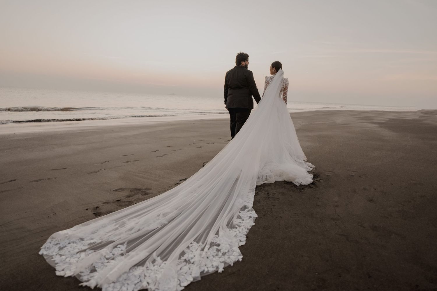 A couple walks along a beach at sunset with the bride's long lace train flowing behind them.