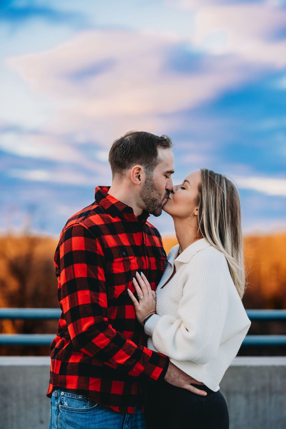 A couple shares a romantic kiss while wearing winter attire against a sunset sky.