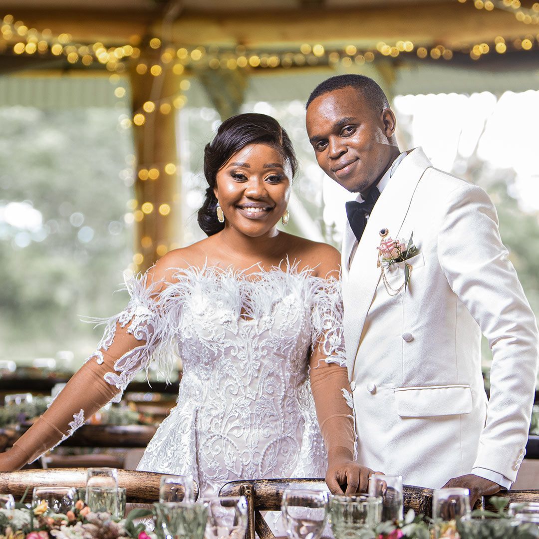 Newlyweds stand together at their reception table decorated with twinkling lights and elegant decor.