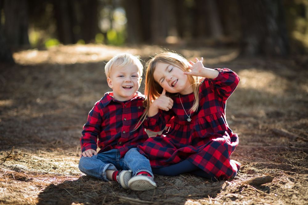 Two children in matching red plaid outfits sitting on ground making silly faces in forest setting.