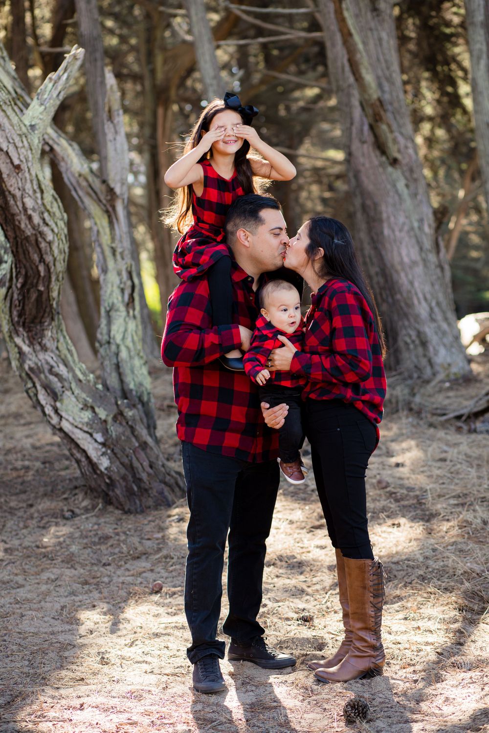 Family wearing matching buffalo plaid shirts sharing a tender moment in a forest setting.