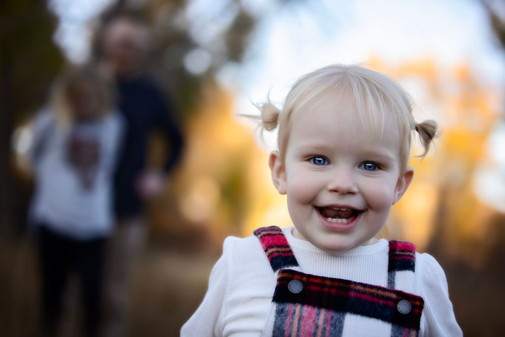 A toddler in plaid overalls smiles brightly with blurred autumn background.