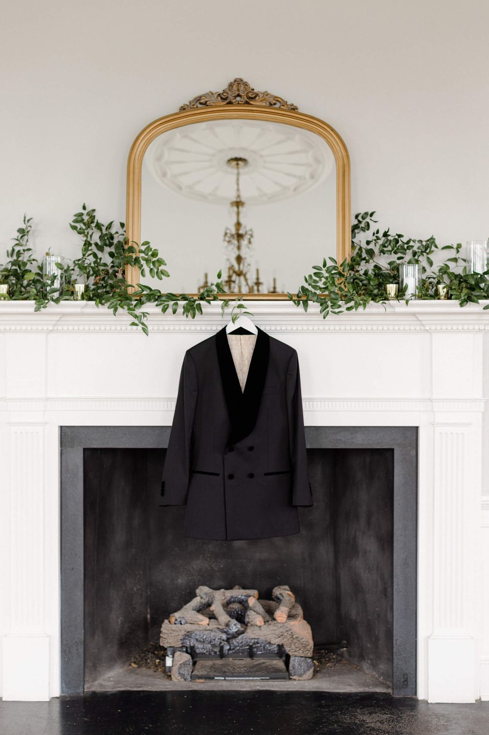 A classic black tuxedo jacket hanging above a white fireplace mantel decorated with greenery.