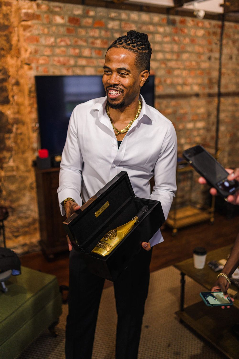 A person in a white dress shirt holds a black presentation box while smiling warmly against a brick wall background.