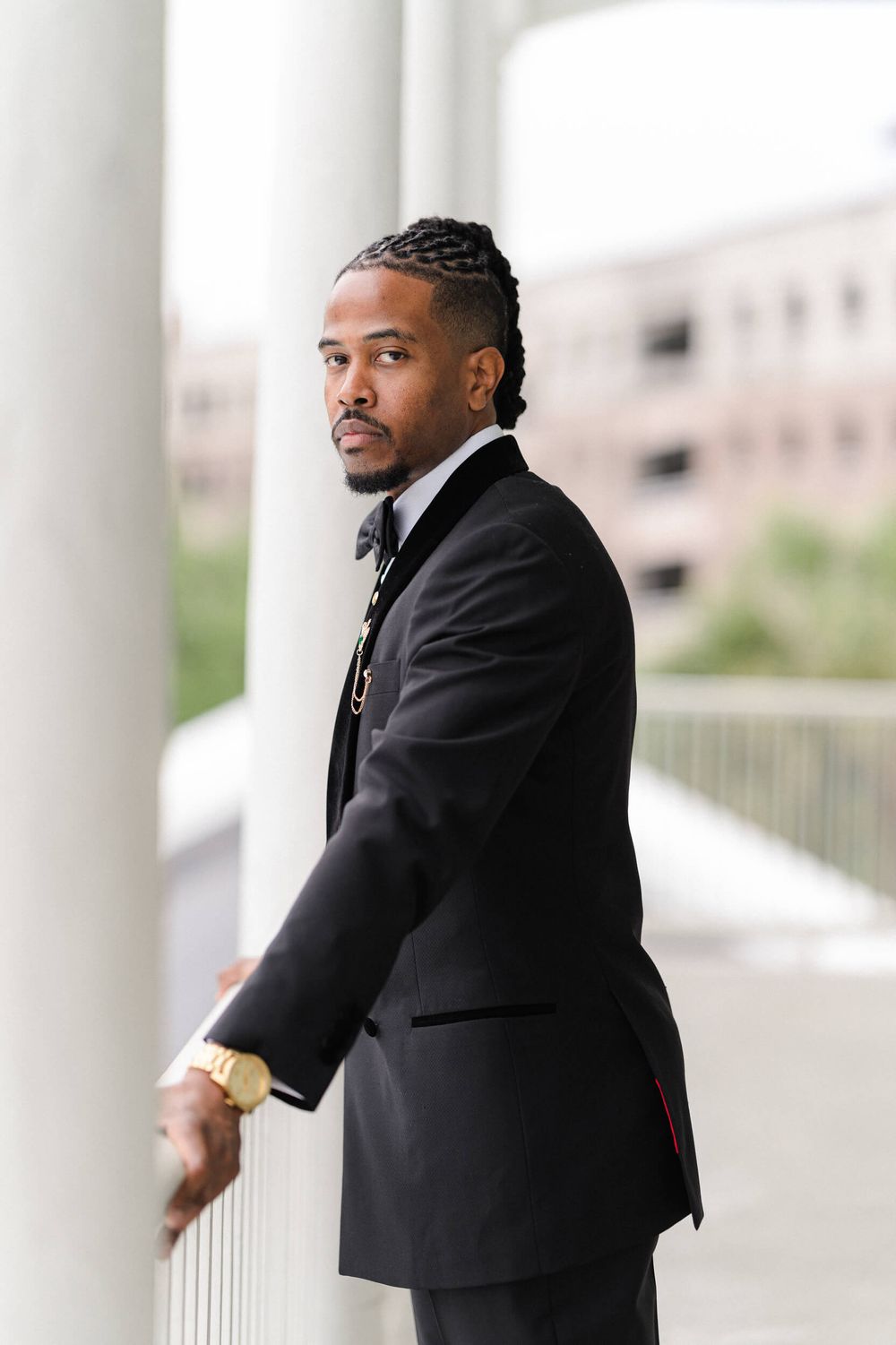 A person in a classic black tuxedo poses thoughtfully against white pillars outdoors.