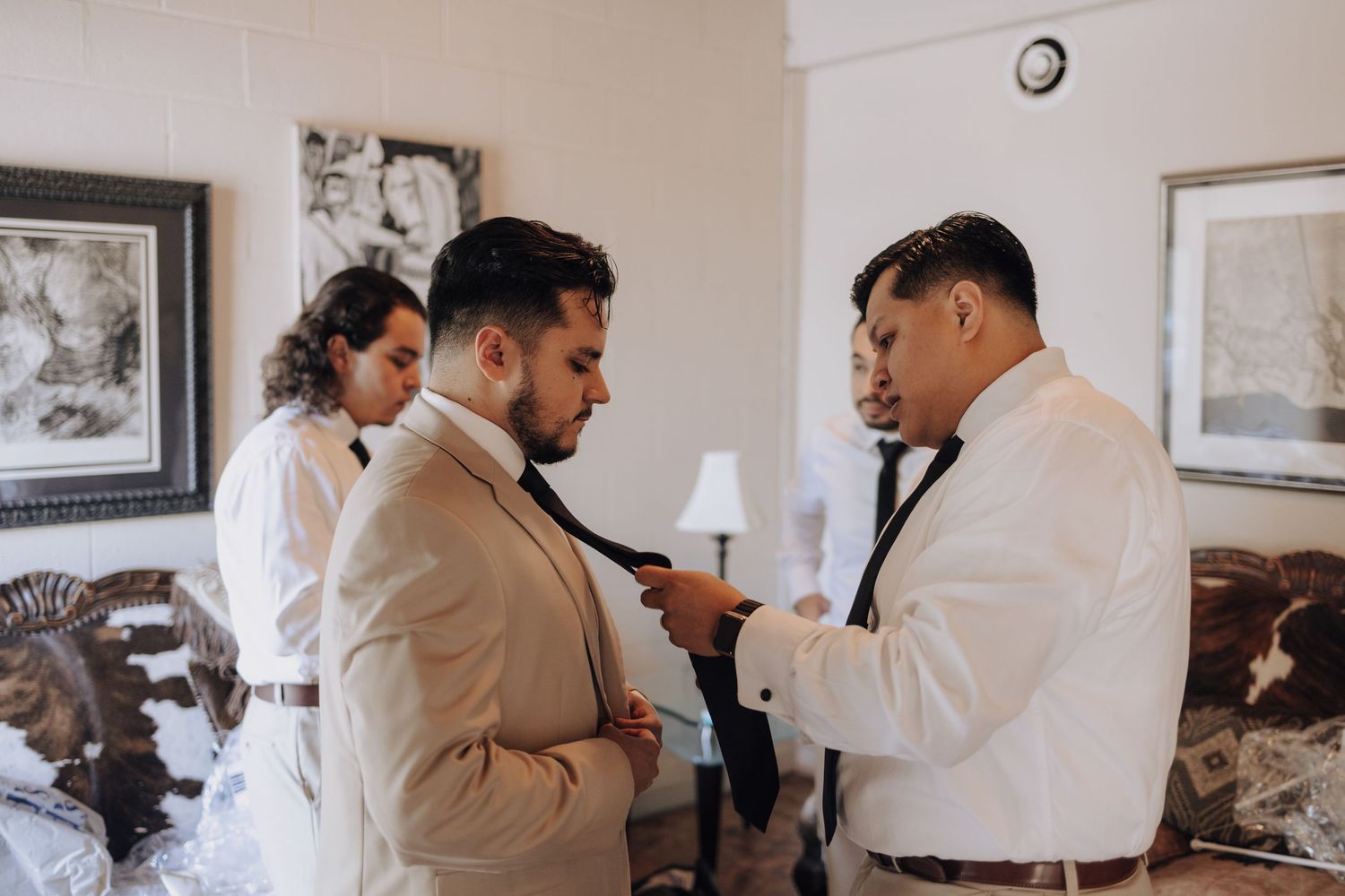 People in formal attire help adjust a tie during wedding preparations.
