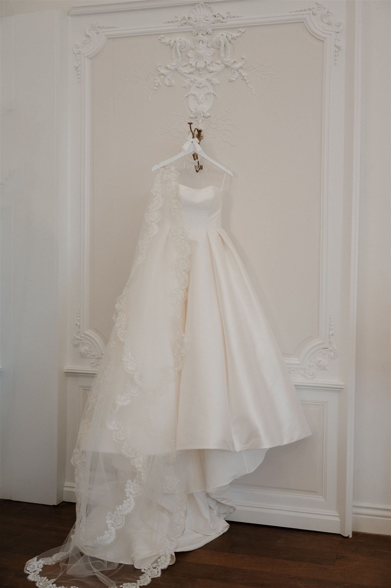 A luxurious white wedding dress with lace details hangs elegantly on a hanger against an ornate white wall with molding.