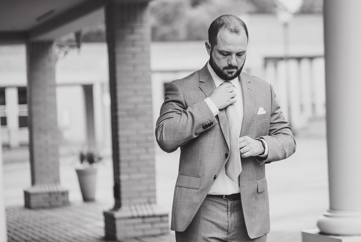 A groom in a light gray suit adjusts his tie while standing on a porch.