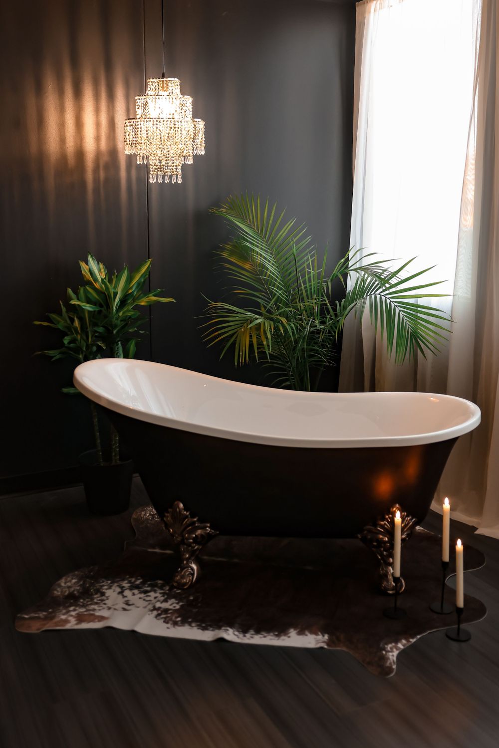 Luxurious clawfoot bathtub with crystal chandelier and tropical plants against dark walls.