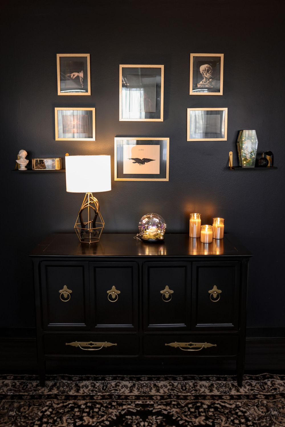 Dark dresser with vintage frames, table lamp, and candles against black wall with ornate decor.