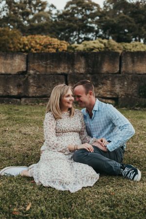 A couple shares an intimate moment while sitting on grass against a stone wall backdrop.