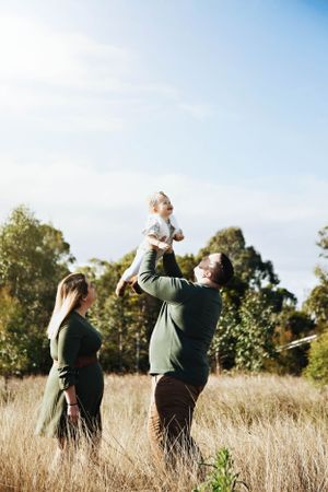 A couple in green sweaters lifts their child high in a sunlit meadow with trees in the background.
