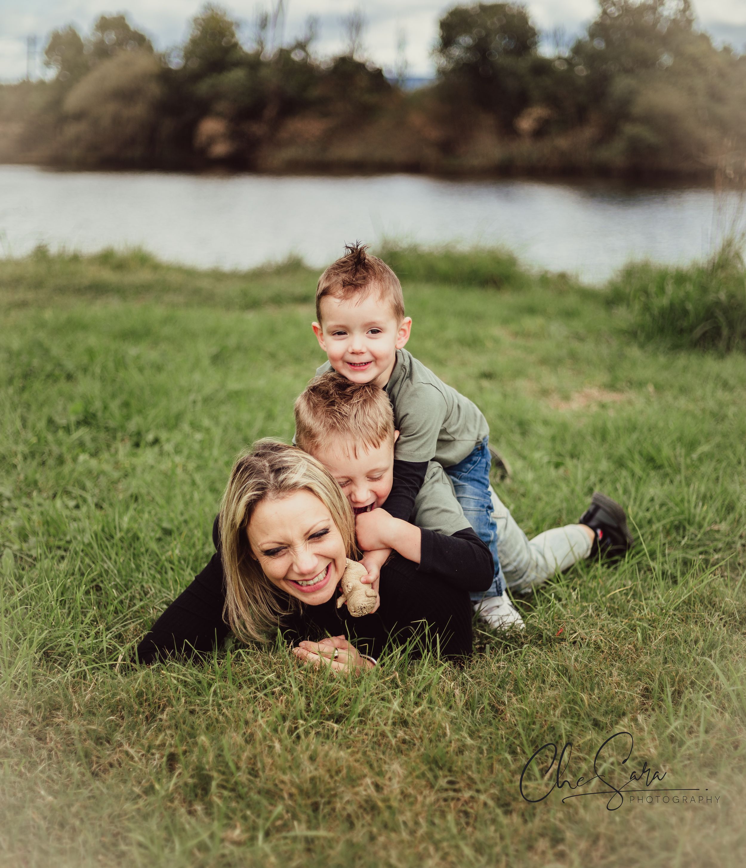 A playful moment captured on grass by a lake as family members pile on top of each other laughing.