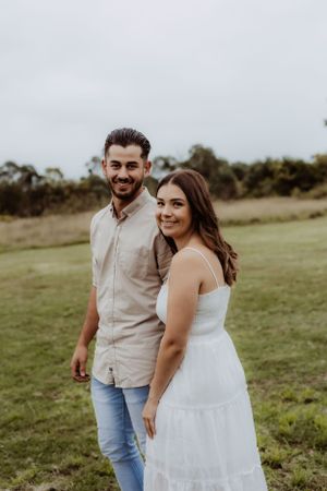 Couple poses together in casual attire against green field backdrop during outdoor photoshoot.
