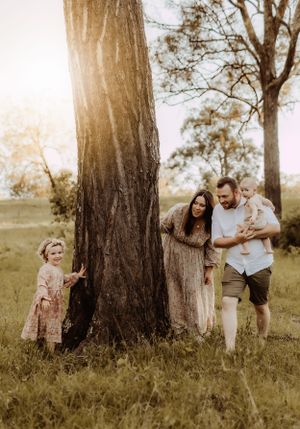 Family plays hide and seek around large tree trunk during sunset photo session.