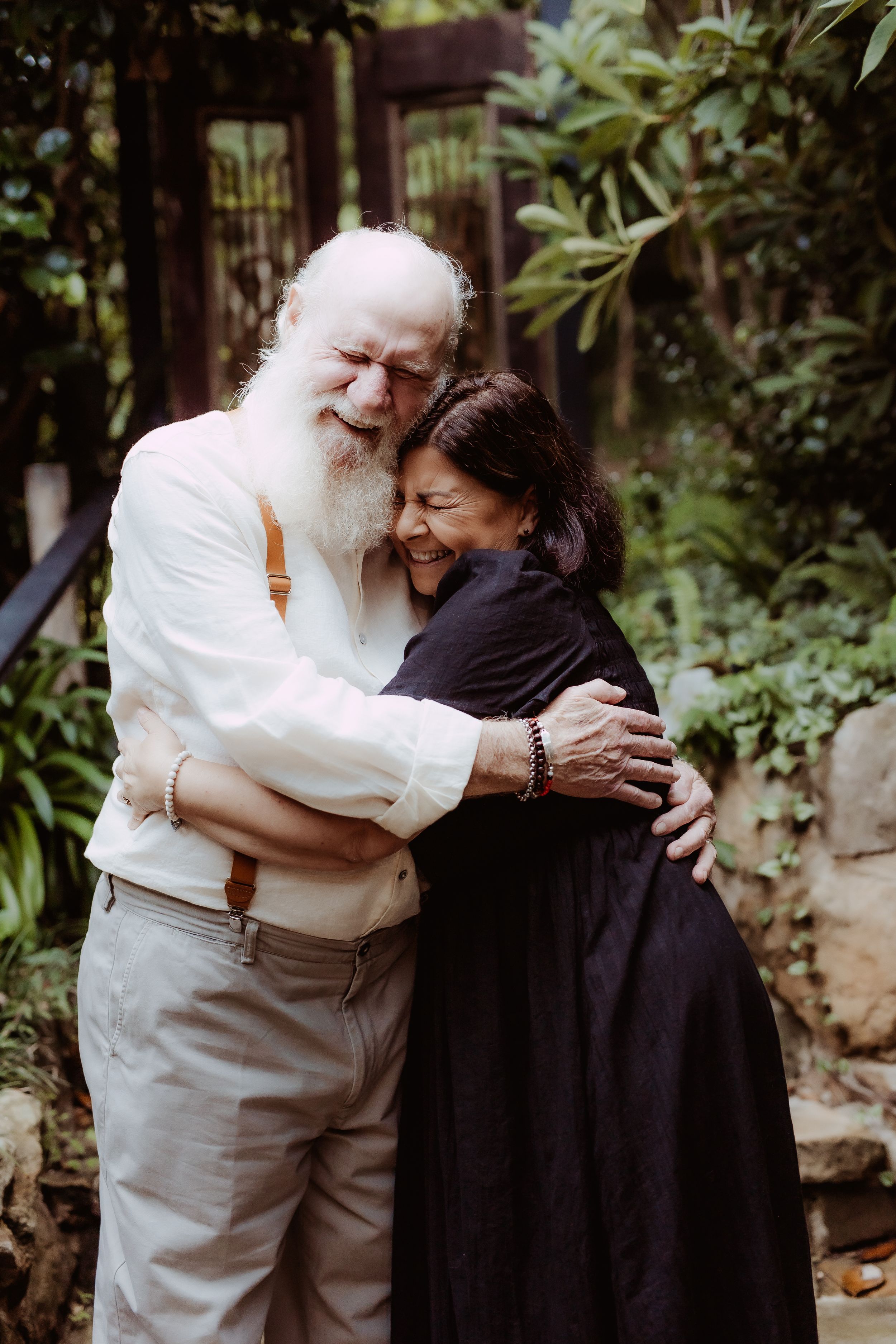 Two people share a warm embrace in a garden setting with lush greenery in the background.