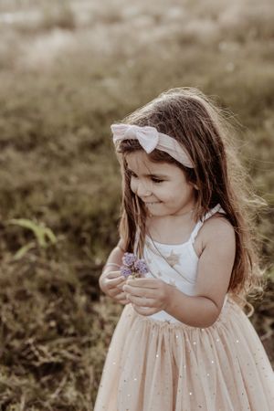 Young child in a sparkly peach dress examines something in their hands outdoors.