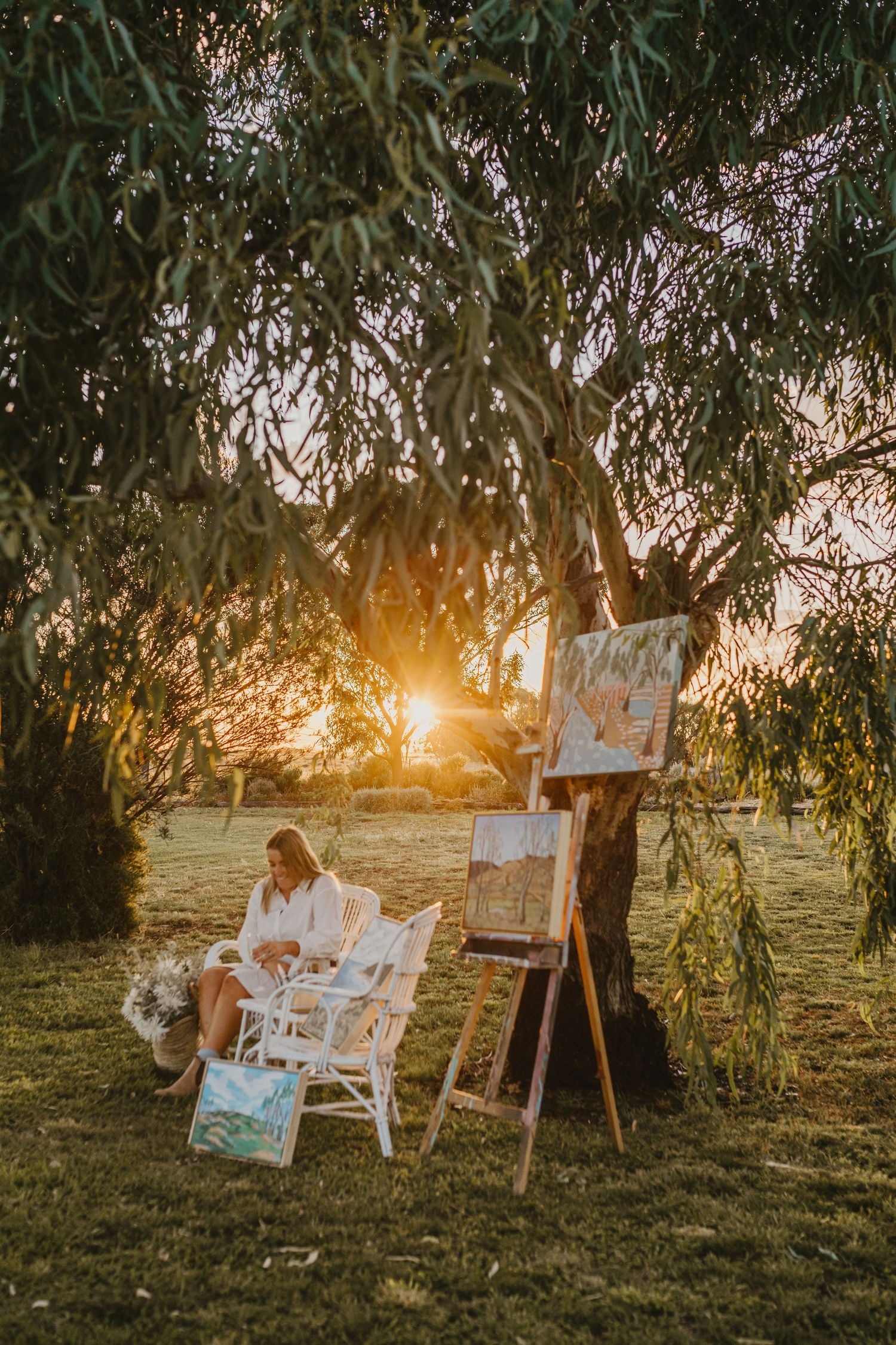 Molly Deans rural artist relaxing in a white chair in her rural garden with her art around her at sunset under a tree