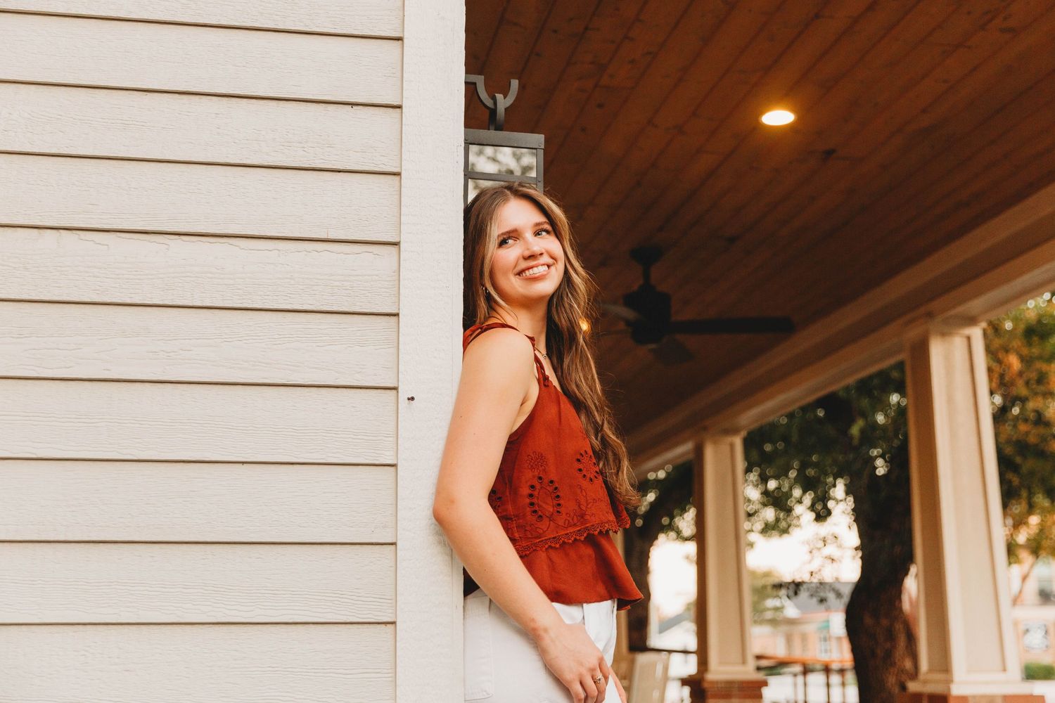 Senior irl in red sleeveless top poses against white porch column during golden hour.
