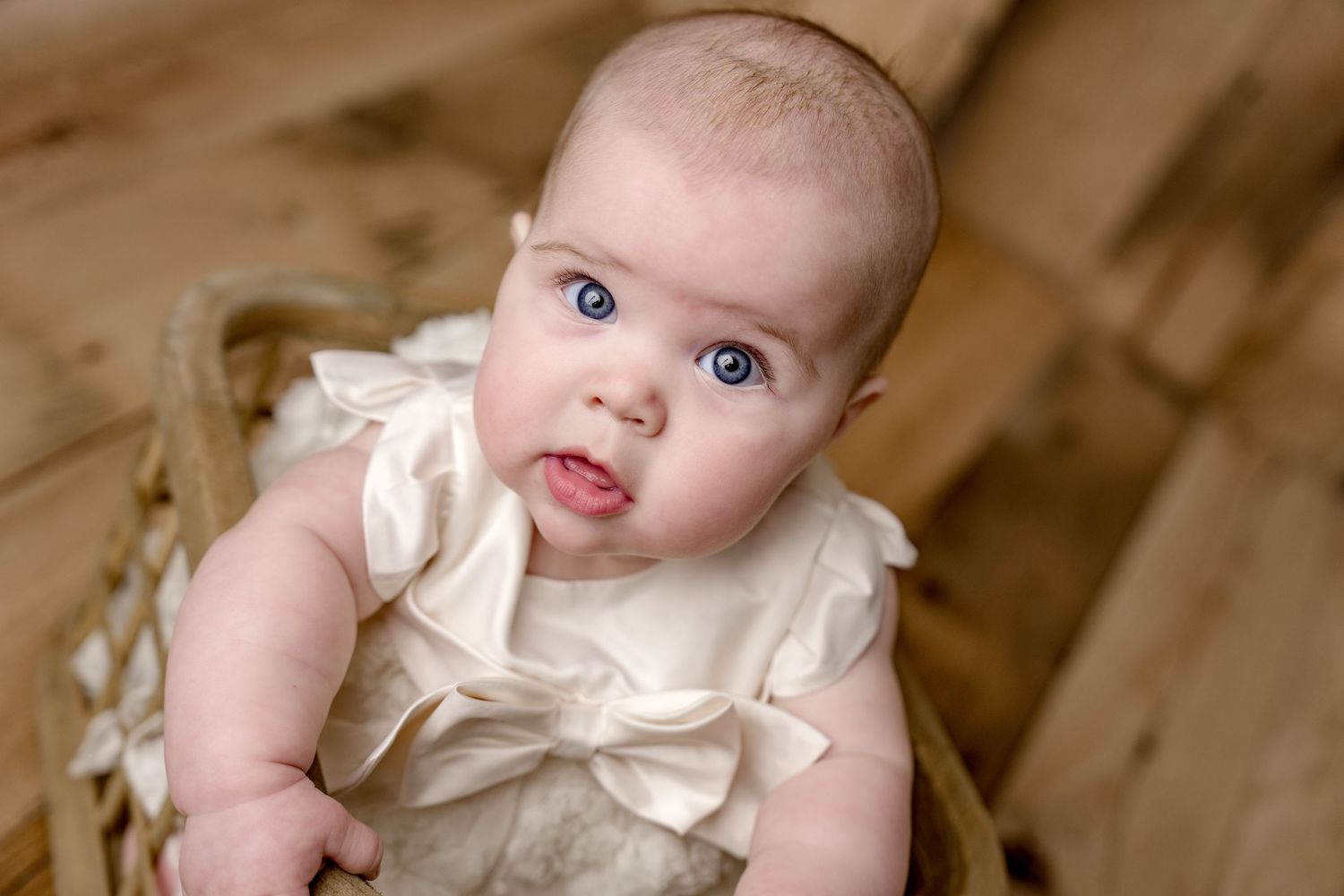 Baby wearing white dress with bow detail photographed on wooden background.