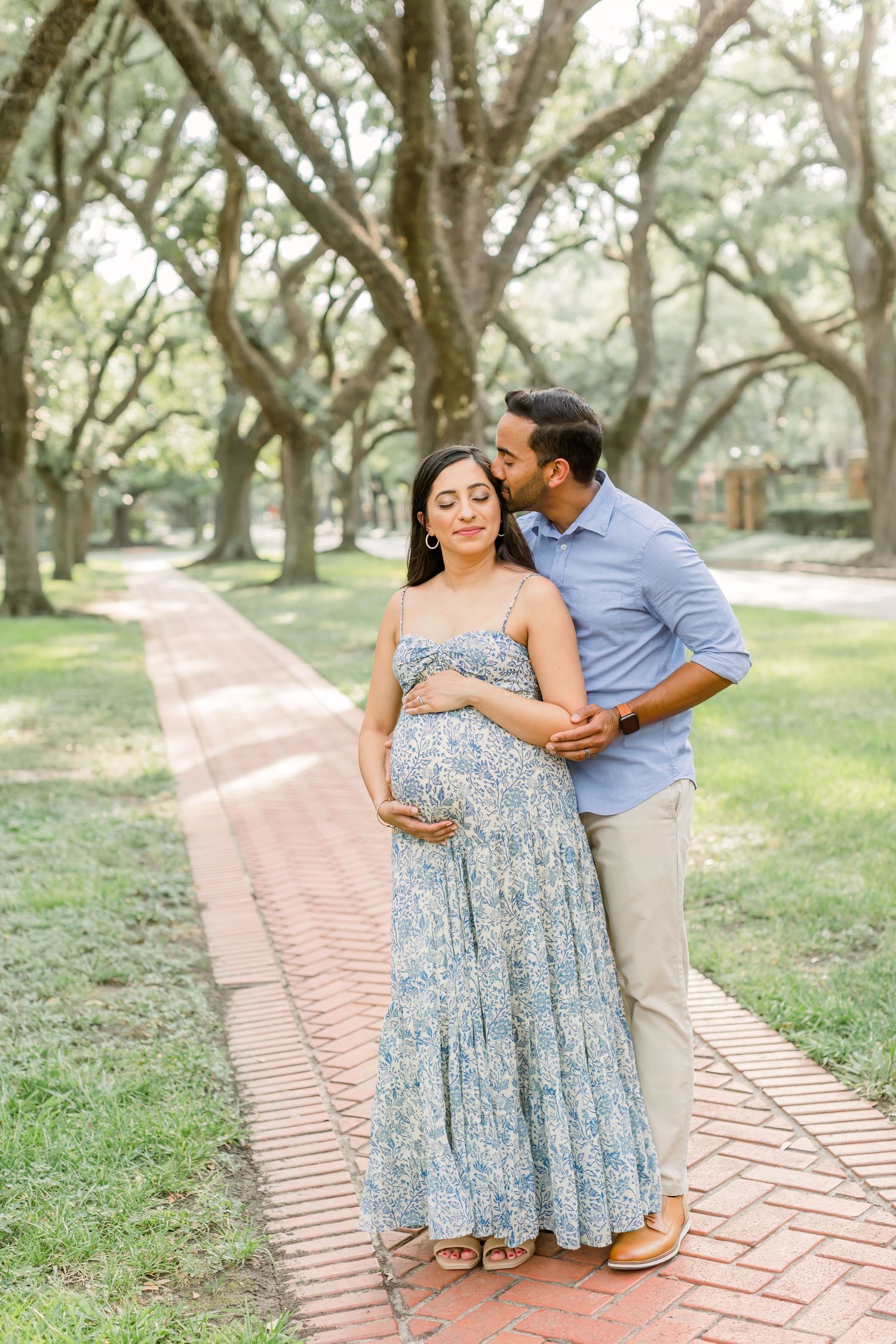 Loving couple poses for maternity photos on brick pathway under canopy of oak trees.