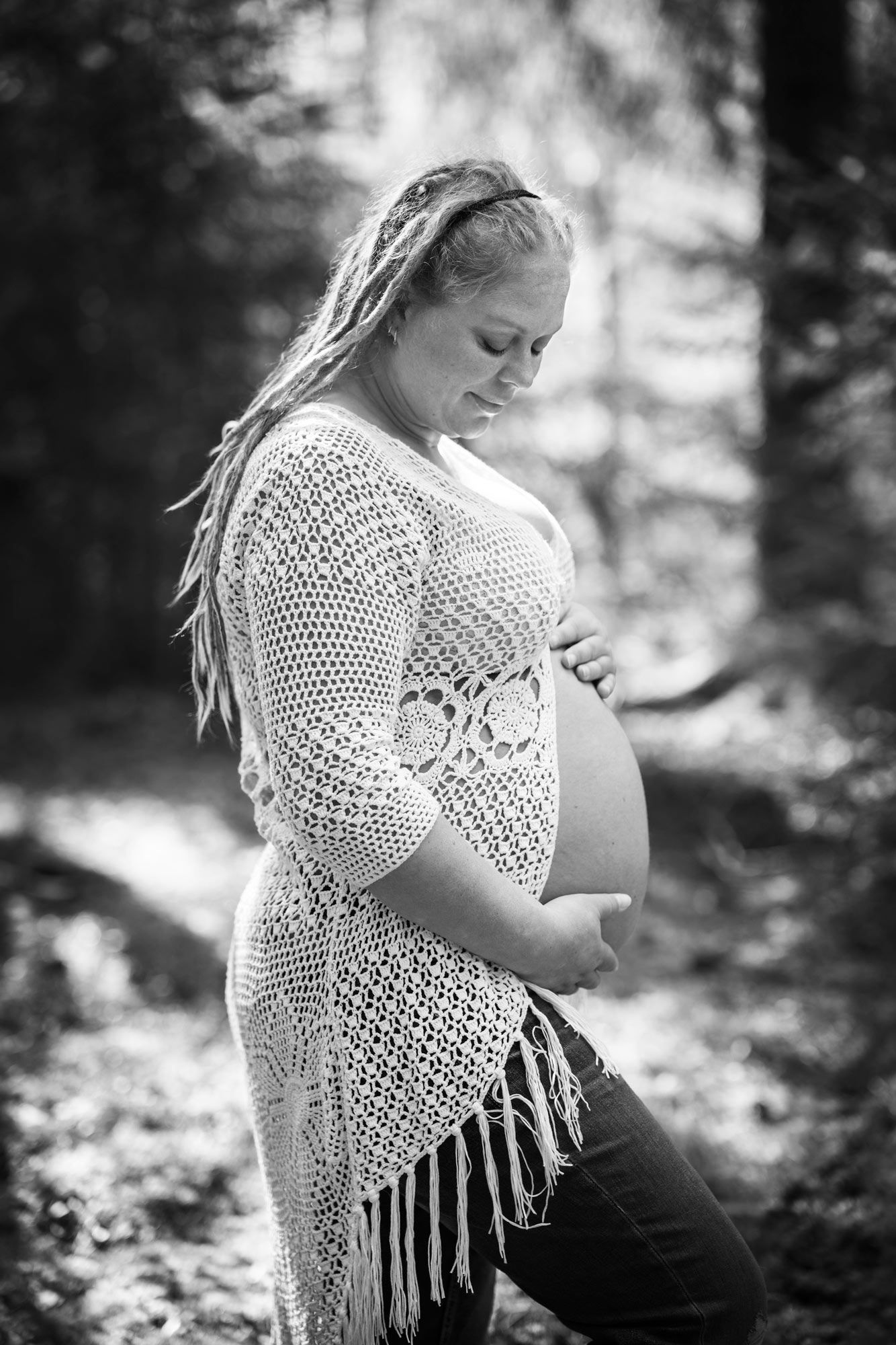 En svartvit bild på en blond kvinna med långa dreadlocks i virkad kofta håller om sin gravidmage. Hon står i skogen. Det är solig.