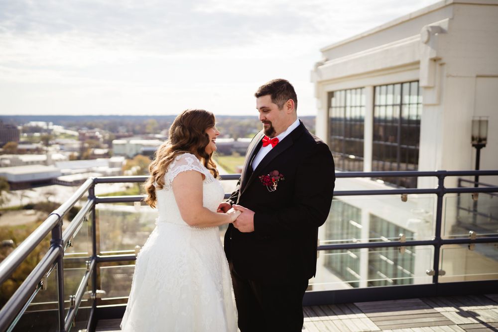 A couple shares an intimate moment in wedding attire on a high-rise balcony with city views.