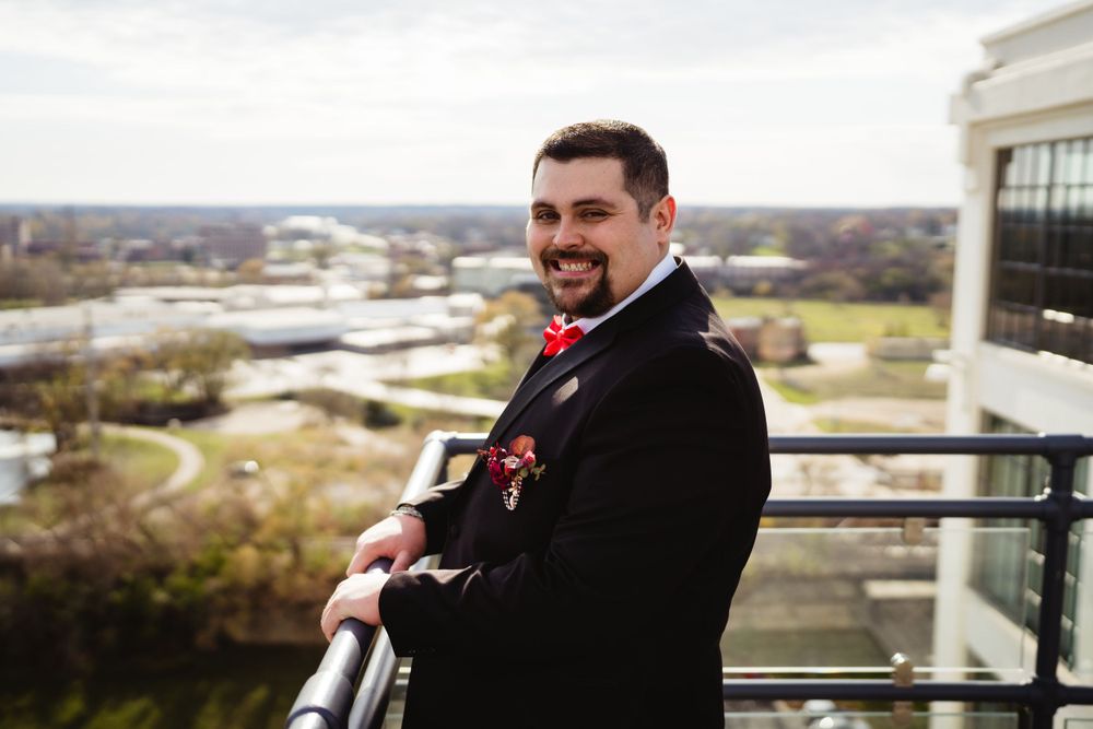 A smiling person in formal attire poses on a balcony overlooking a city with a river in the background.