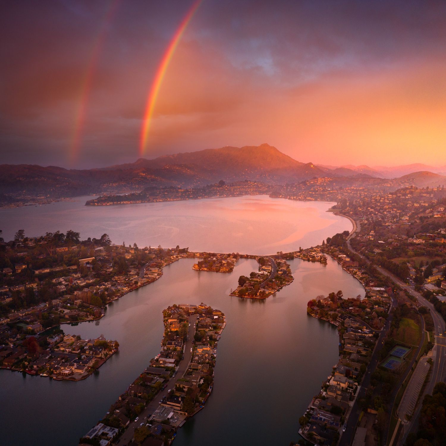 Double rainbow arches over Marin County and mountains at sunset with reflective waters and residential docks.