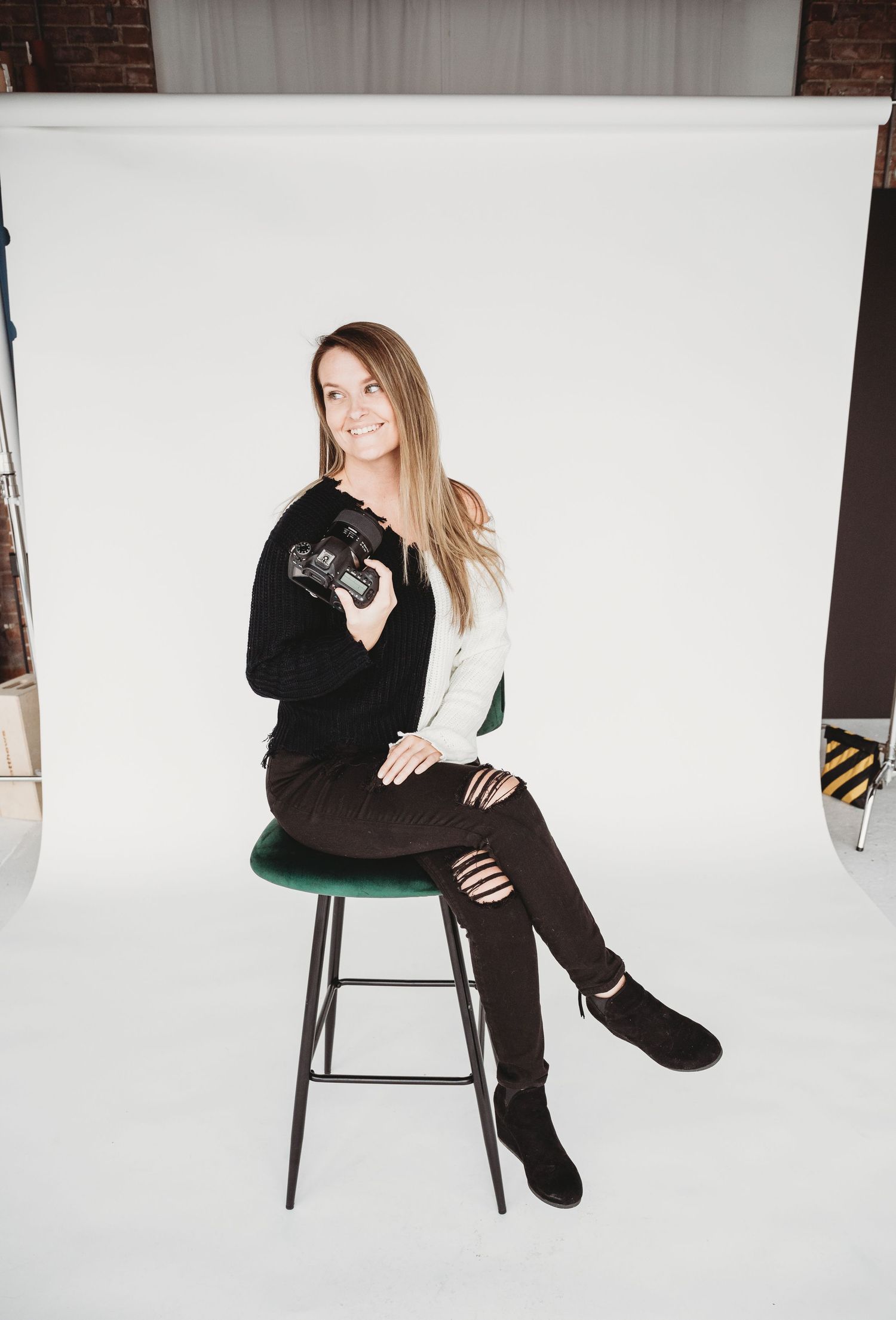 Model in black outfit poses on white stool against white backdrop in studio photography series.