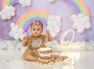 Baby in gold outfit celebrating first birthday with cake against rainbow and cloud backdrop.