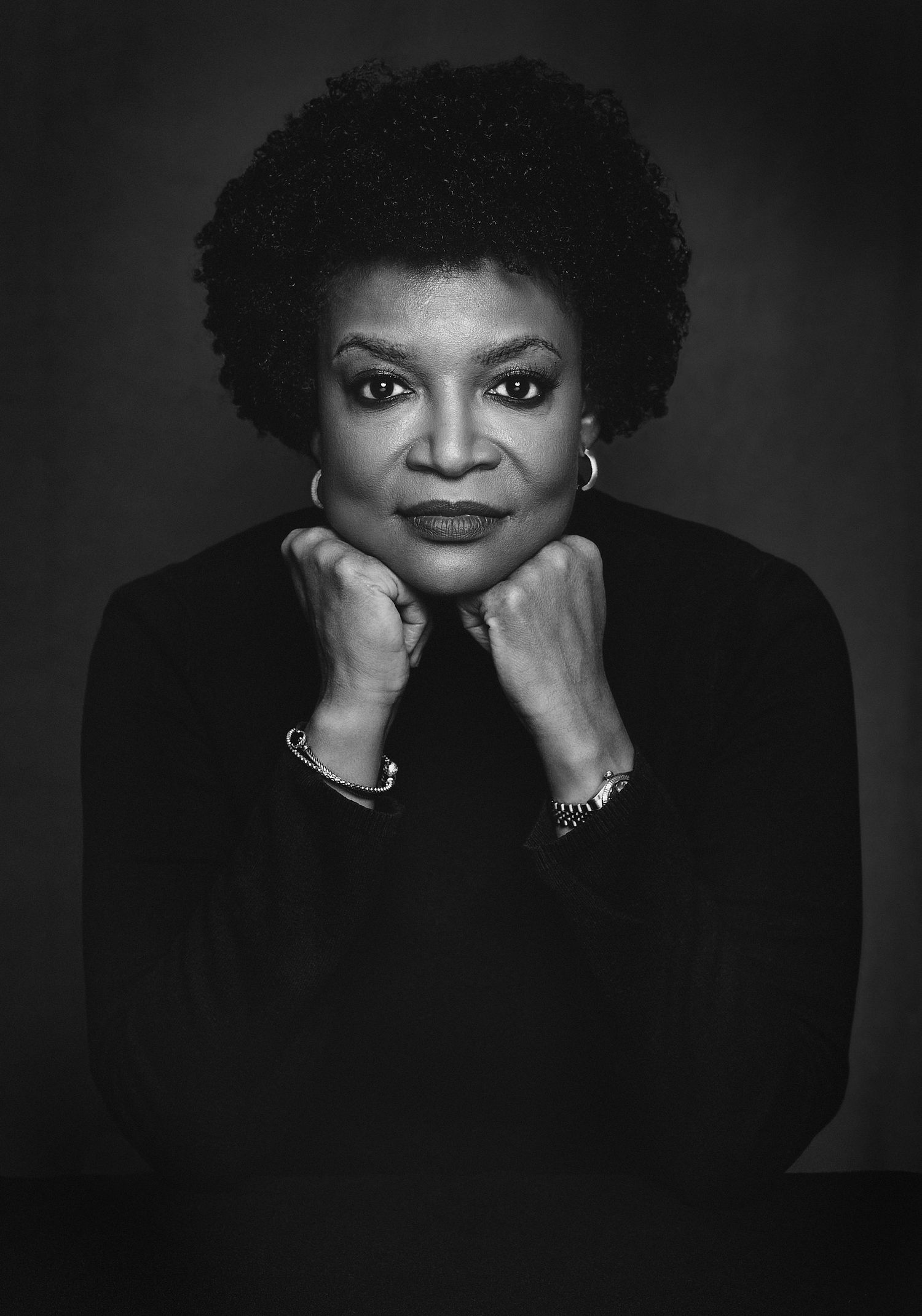 Elegant Black woman with natural afro resting chin on clasped hands in dramatic black and white portrait.