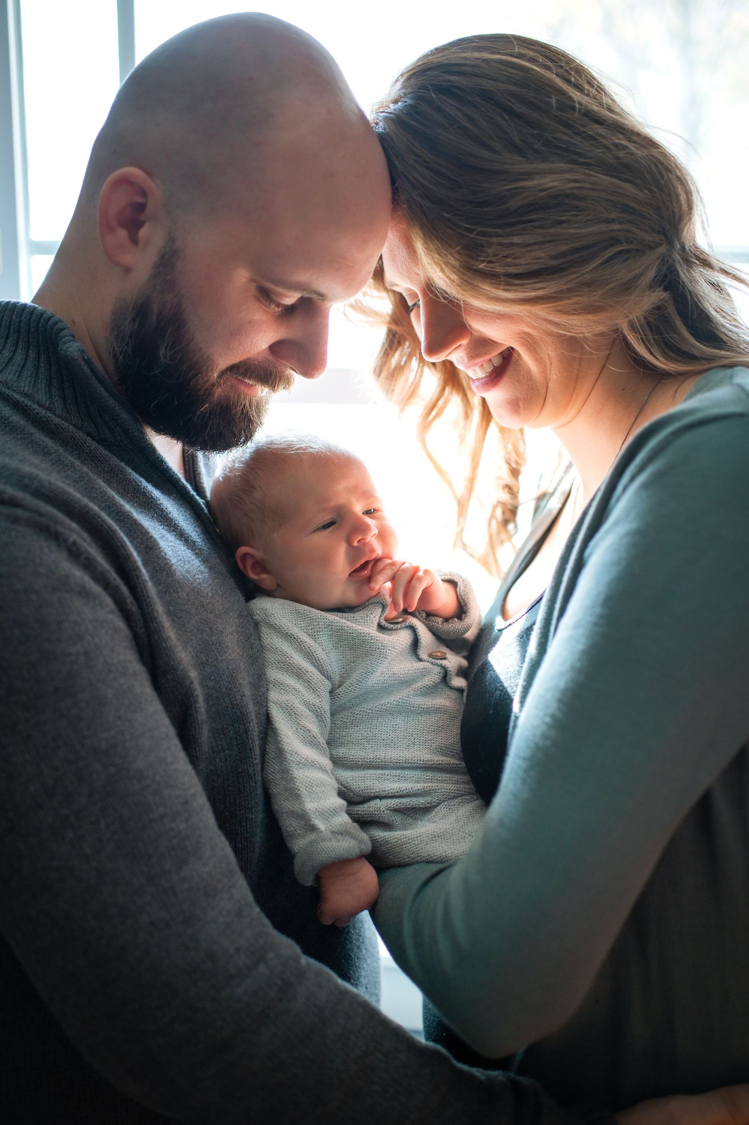 Young family shares an intimate moment with their newborn baby in soft window light.