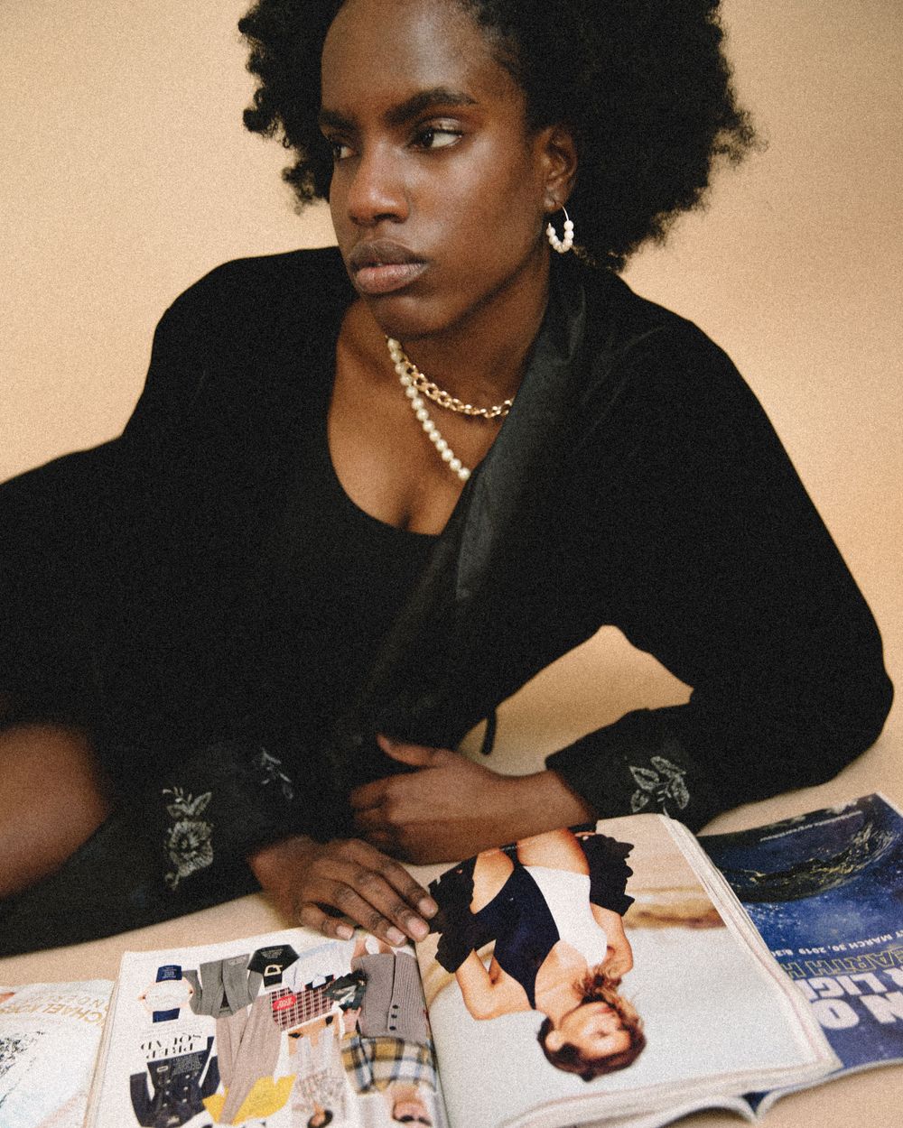 Fashion editorial of a model in black with pearl necklace looking at magazines against beige background.