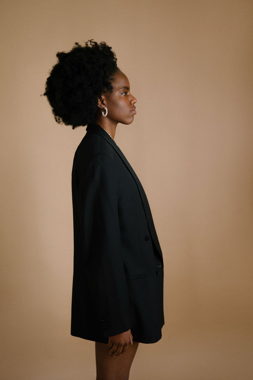 Minimalist fashion portrait featuring black blazer against warm neutral background.