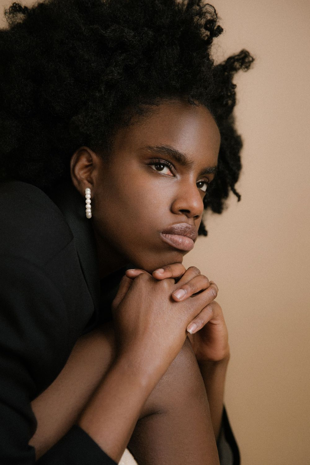 Contemplative portrait with pearl drop earrings and black blazer against neutral background.