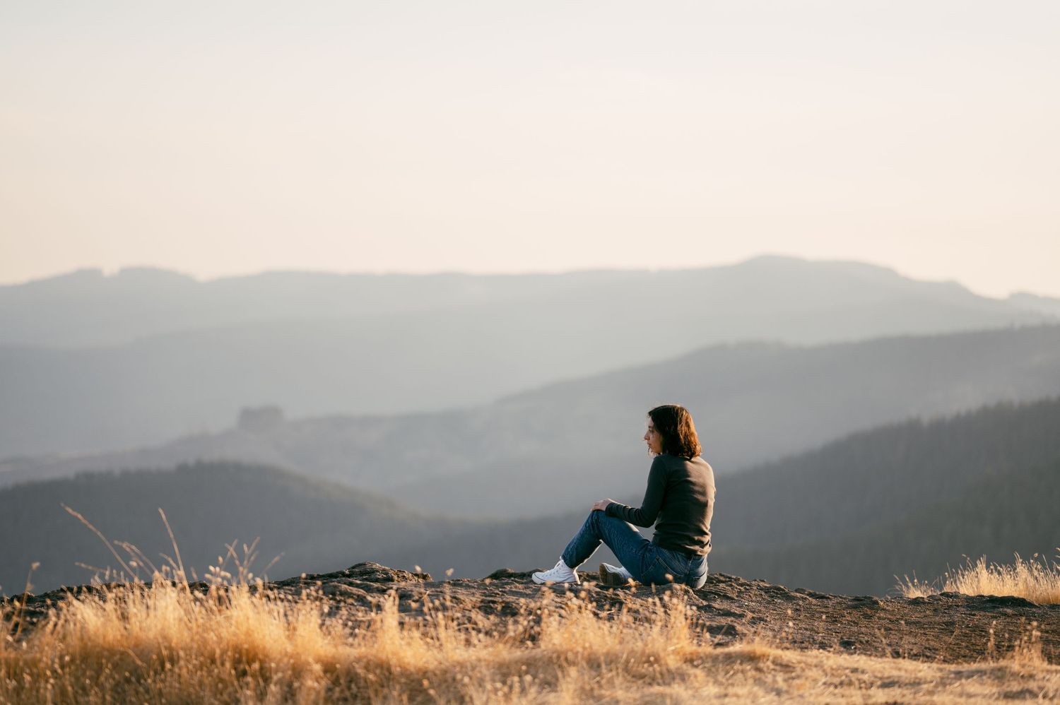 A silhouetted figure sits peacefully on a hilltop overlooking mountain ranges at sunset.