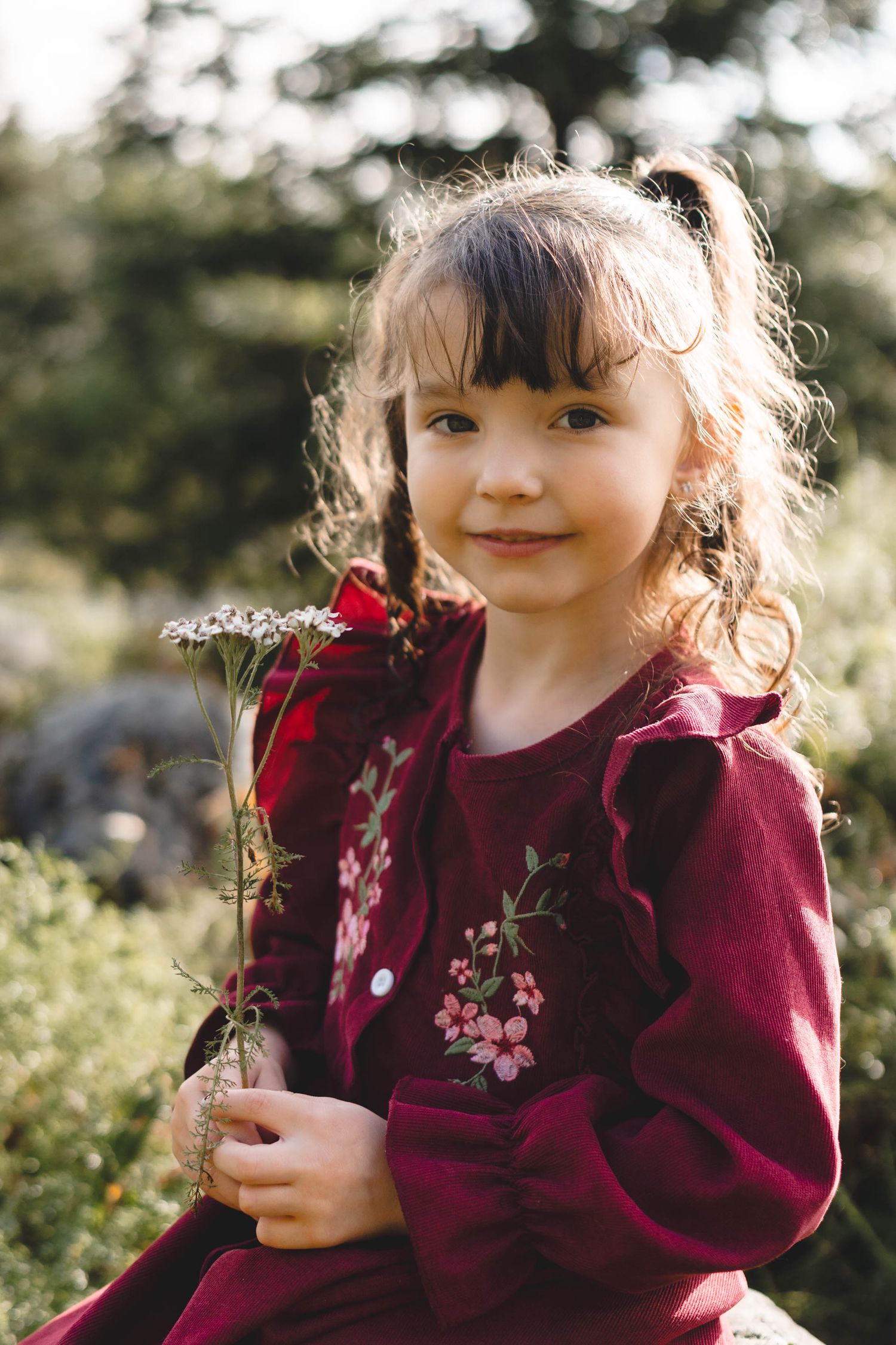 Adorable little girl in a red floral dress holding wildflowers while smiling outdoors in nature.
