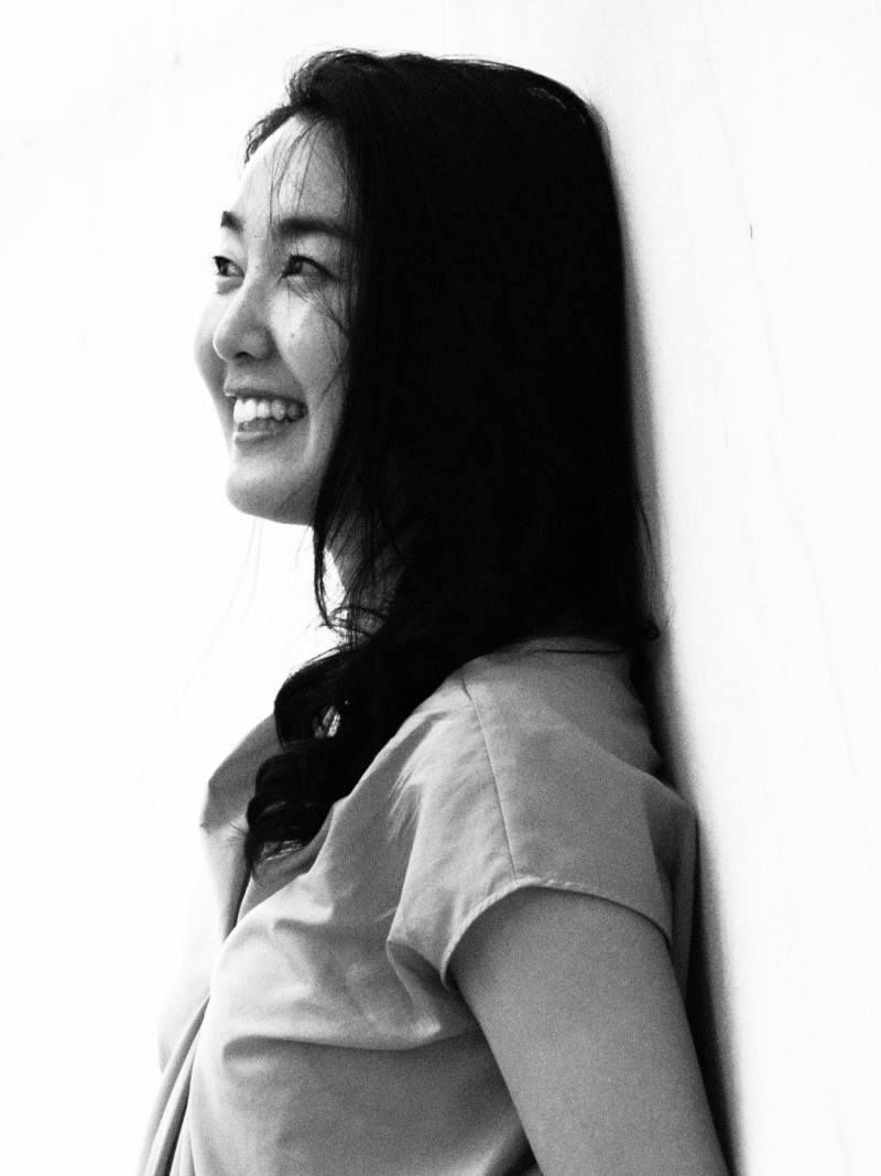 Black and white profile portrait of a smiling young Asian woman with long dark hair leaning against a wall.
