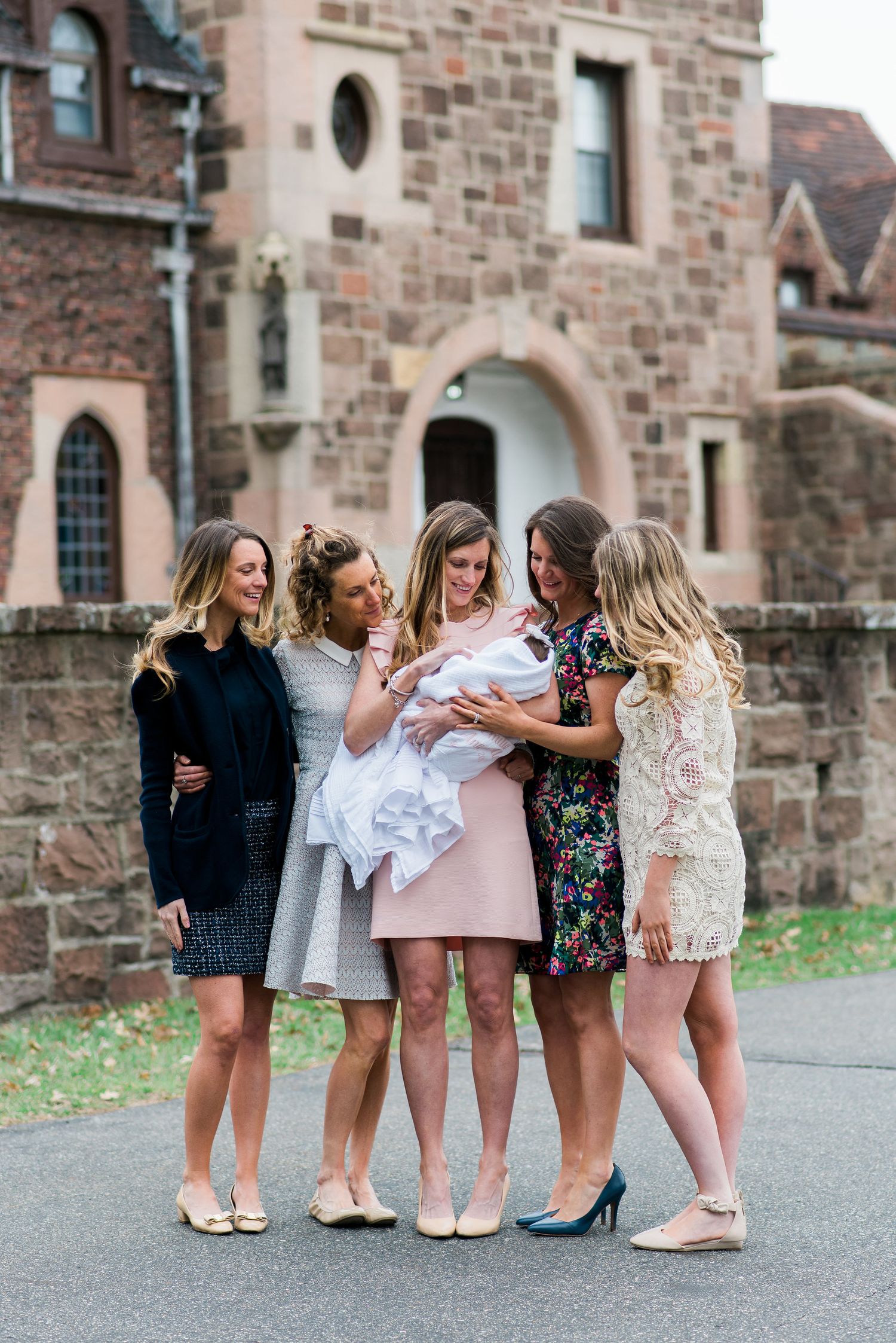 A group of sisters in stylish dresses gather around a baby in white dress outside a historic stone building in West Orange.