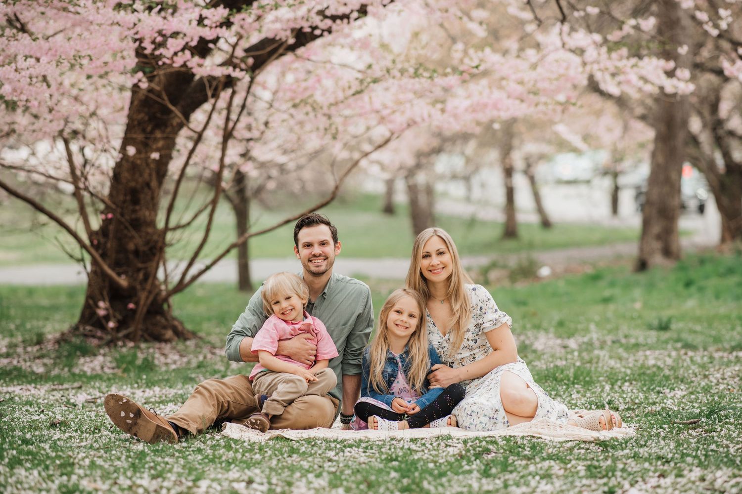 Group sitting on blanket under blooming cherry blossom trees in a spring garden setting.