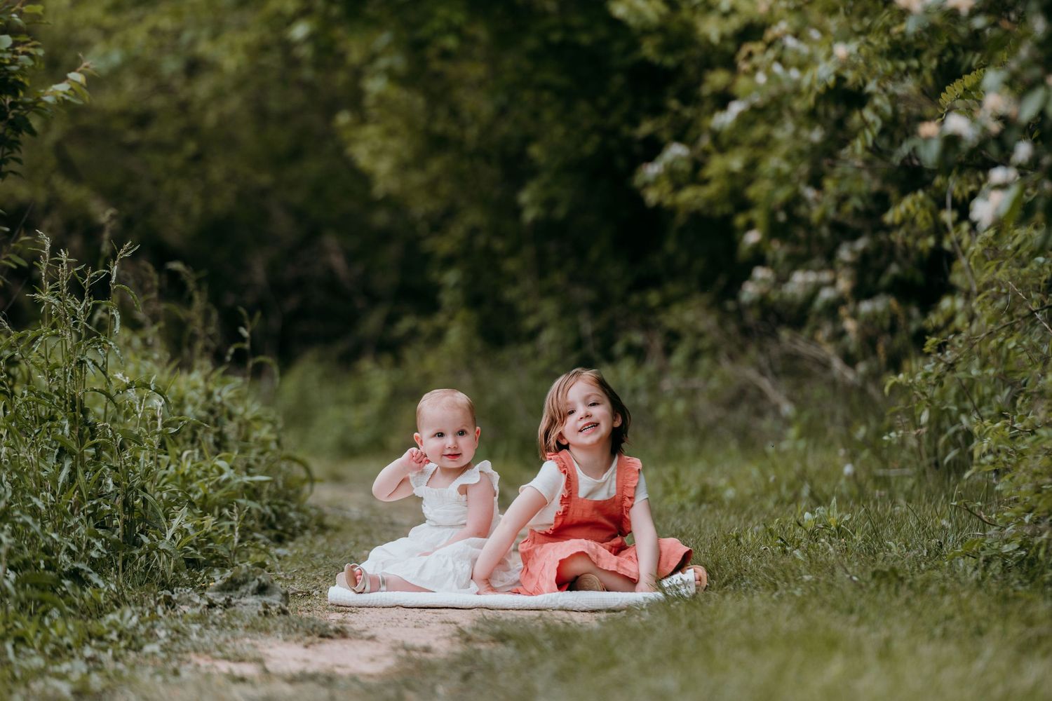 Two children sit together on a blanket in a lush green path surrounded by summer foliage in Montclair.